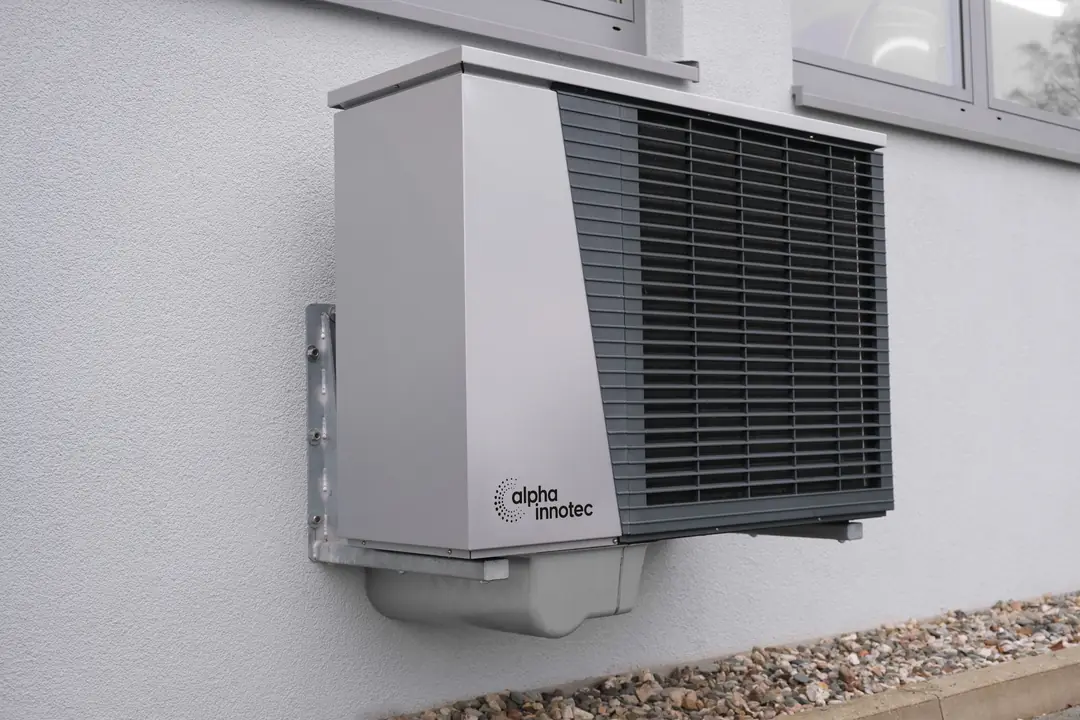 Unité extérieure de pompe à chaleur fixée sur console contre une façade grise, au-dessus d’un lit de graviers